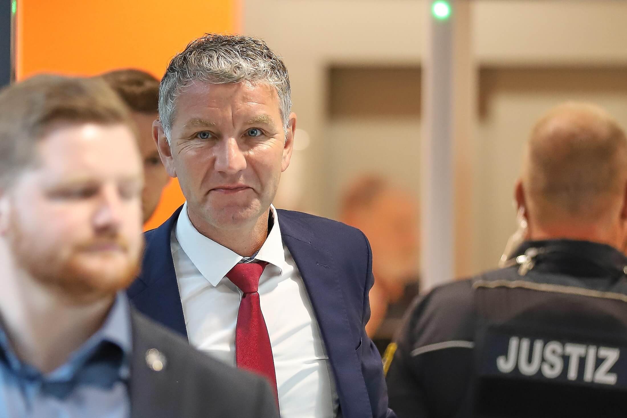 Der AfD-Politiker ist im Fokus des Bilds, er trägt Anzug und Krawatte. Höcke steht im Amtsgericht Halle. Vor ihm läuft ein weiterer Mann, neben ihm steht eine Person in Uniform mit der Aufschrift „Justiz".
